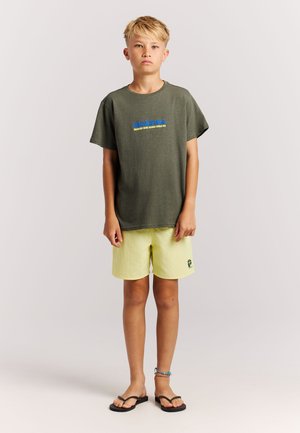 Olivengroen katoenen T-shirt met blauwe en gele tekst, gecombineerd met lichtgele korte broek met een logo. Zwarte slippers maken de outfit compleet.