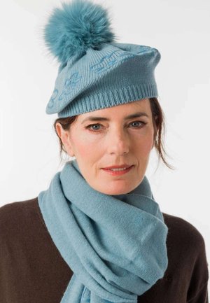 Donna che indossa un cappello di lana azzurro chiaro con pon pon e sciarpa coordinata, vestita con una maglia marrone scuro, con espressione facciale neutra.