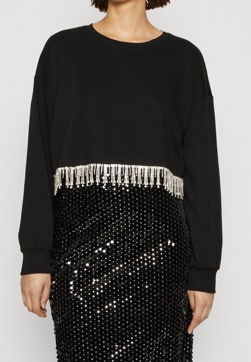 Sweat-shirt noir à épaules tombantes, avec un ourlet à franges perlées. Associé à une jupe noire à paillettes, reflétant la lumière en motifs.
