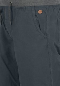 Shorts gris foncé en tissu léger, présentant une texture douce, des poches latérales et un petit label circulaire de la marque sur la ceinture.