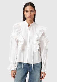 Kvinna med rakt mörkt hår som bär en vit blus med volanger och jeans i blått, stående mot en enkel ljus bakgrund.