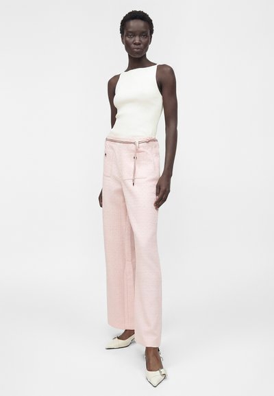 Γυναίκα που φοράει λευκό ασύμμετρο αμάνικο τοπ, ροζ ανοιχτόχρωμο παντελόνι με φαρδιά μπατζάκια και τσέπες, και λευκά μυτερά παπούτσια με αγκράφες.