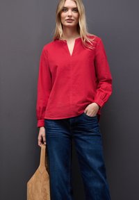 Camisa roja de manga larga con escote en V y puños fruncidos, combinada con pantalones vaqueros azul oscuro y un bolso de ante beige. Color sólido, tejido texturizado.