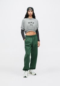 Γκρι cropped φούτερ με μαύρο λογότυπο "NIKE 1972", συνδυασμένο με πράσινη φόρμα και λευκά αθλητικά παπούτσια με πράσινες λεπτομέρειες. Μαύρο καπέλο που φοριέται ανάποδα.