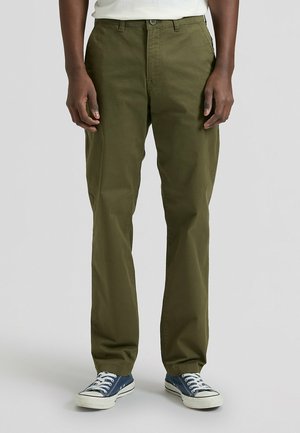 Mężczyzna ubrany w oliwkowozielone spodnie o prostych nogawkach i niebieskie tenisówki z płótna, stojący na tle jednolitego jasnoszarego tła.