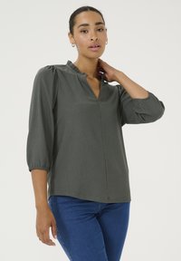 Dunkelgrüne Bluse mit V-Ausschnitt und gerafftem Kragen, ausgestattet mit Dreiviertelärmeln und einem dezenten strukturierten Stoff. Kombiniert mit blauen Jeans.