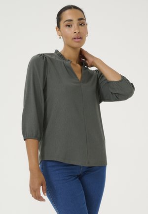 Mørkegrøn bluse med v-udskæring og flæsekrave, med trekvartærmer og en subtil struktureret stof. Kombineret med blå jeans.