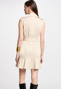 Beige mouwloze jurk met schouderepauletten, getailleerde taille, uitlopende zoom en strik aan de achterkant. Gecombineerd met een gouden armband en kniehoge zwarte laarzen.