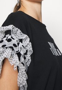 Černé tričko s volány na rukávech vyrobené z průhledné bílé látky s černými puntíky; text "VÍCE VOLNOČASU" bílou barvou na přední straně.