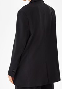 Svart skräddarsydd kavaj med en mjuk textur, figursydd silhuett och slagkrage. Har knäppte manschetter och en slits bak.