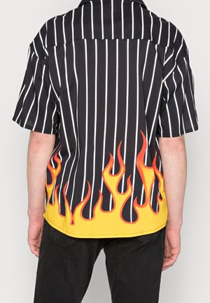 Set bagfra af person iført sort kortærmet skjorte med hvide lodrette striber og gul flammedesign i nederste kant.