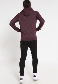 Sudadera color burdeos con logo en la espalda, capucha y puños acanalados, combinada con pantalones de chándal negros y zapatillas blancas con marca negra.
