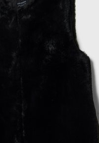 Veste sans manches en fausse fourrure noire avec col rond, étiquette de marque visible au col, texture lisse et bords droits.