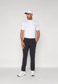 Hvit poloshirt med krage og logo, kombinert med svarte bukser og hvite joggesko. Modellen har også på seg en hvit caps, stående foran en nøytral bakgrunn.