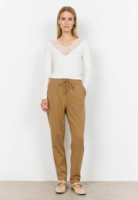 Weißes Langarmoberteil mit Spitzen-V-Ausschnitt, kombiniert mit hellbraunen Jogginghosen mit Kordelzug. Das Modell trägt beiges Slipper. Glatte Textur.