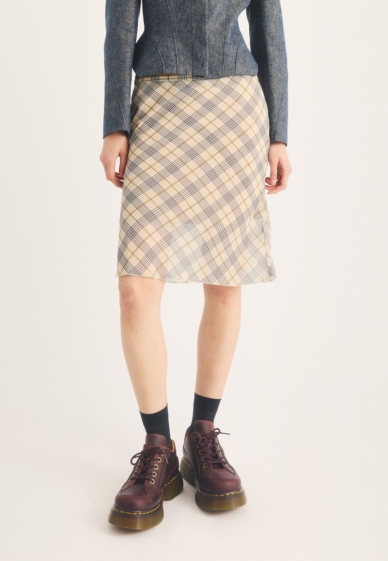 Femme portant une jupe beige à carreaux, une veste grise, des chaussettes noires et des chaussures à lacets épaisses rouge foncé, debout sur un fond blanc.
