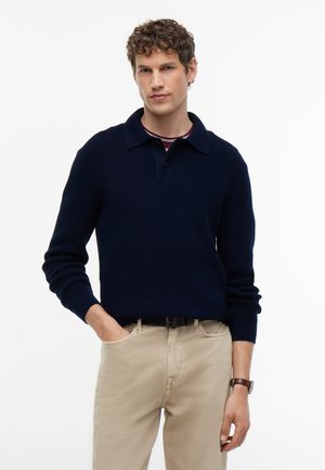 Homme aux cheveux bouclés portant un pull bleu foncé et un pantalon beige, debout avec une main dans la poche devant un fond blanc uni.