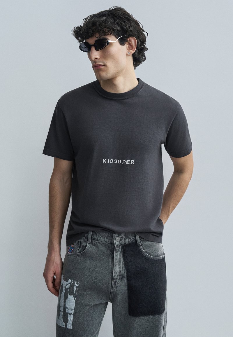 Σκούρο γκρι βαμβακερό t-shirt με το κείμενο "KIDSUPER" τυπωμένο σε λευκό, σε συνδυασμό με φθαρμένο γκρι τζιν με σχέδια σε μπαλώματα.