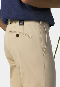 Beige Hosen mit einer strukturierten Oberfläche, ausgestattet mit einer Gesäßtasche mit Knopfverschluss und einem gestickten Detail darüber. Kombiniert mit einem marineblauen Hemd.