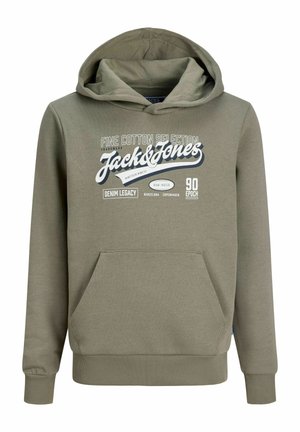 Plavokosi dječak u zelenoj Jack & Jones dukserici s kapuljačom, plavim trapericama i bež cipelama stoji i gleda u stranu na jednobojnoj pozadini.