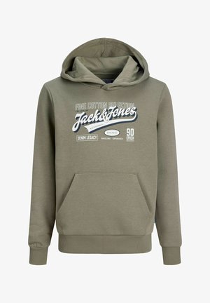 Blond poiss rohelise Jack & Jonesi kapuutsiga dressipluusi, siniste teksade ja beežide jalanõudega, seisab ja vaatab küljele ühevärvilise tausta ees.