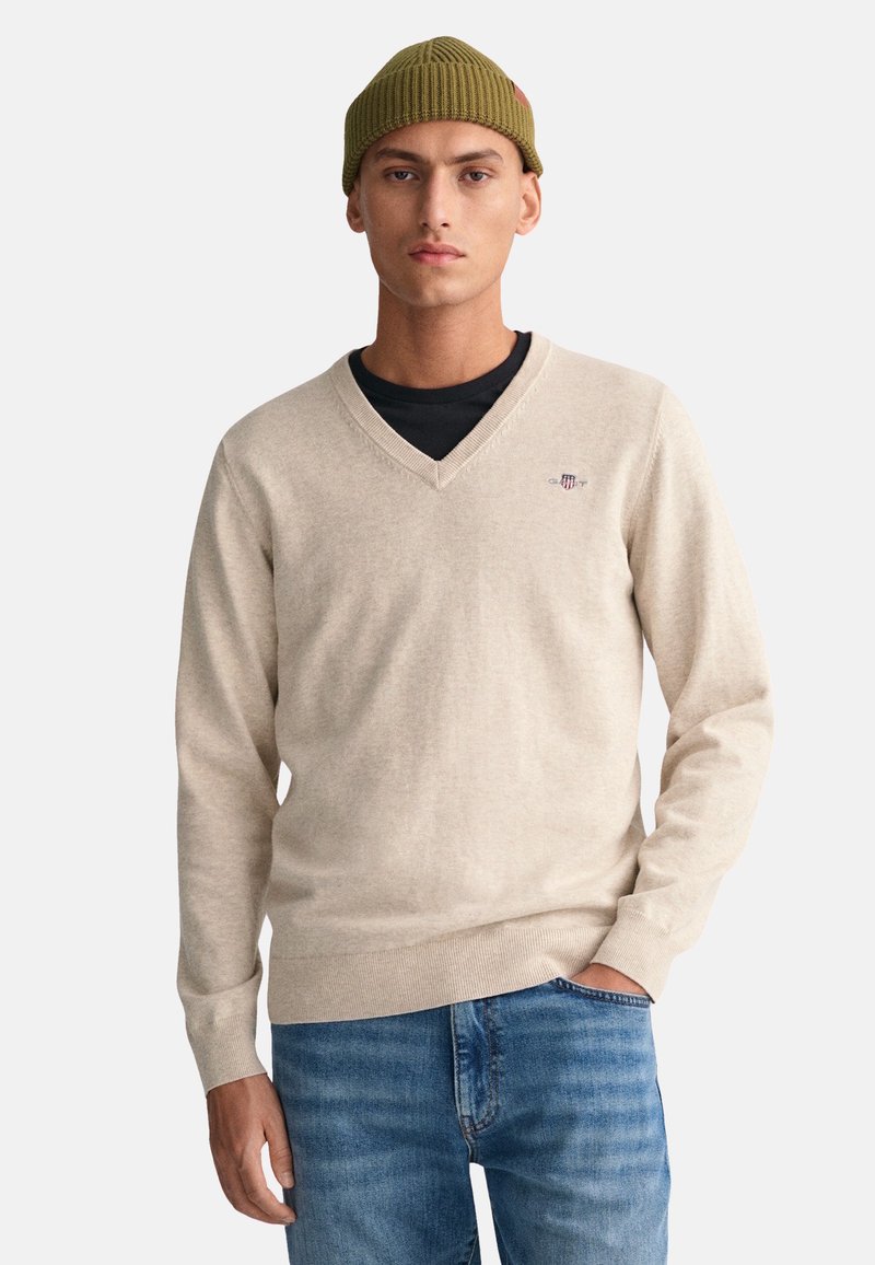 Helles beige V-Ausschnitt-Pullover aus weichem Strickmaterial, mit einem kleinen Logo auf der linken Brust und gerippten Bündchen, kombiniert mit blauen Jeans.