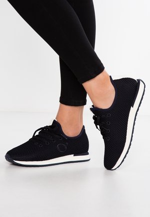 Personne portant des baskets en mesh noir avec des semelles blanches, debout sur une surface blanche avec des leggings noirs.