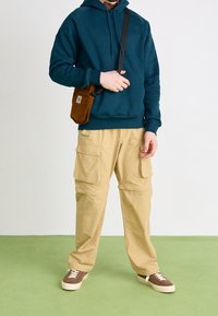 Felpa con cappuccio verde petrolio scuro, pantaloni cargo beige con pi�ù tasche, borsa a tracolla in velluto a coste marrone e scarpe da ginnastica marroni su uno sfondo di pavimento verde.
