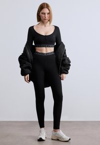 Μαύρο αθλητικό σετ που αποτελείται από τοπ cropped με μακριά μανίκια και κουμπιά και κολάν με ψηλή μέση. Περιλαμβάνει ένα φουσκωτό μαύρο μπουφάν και λευκά αθλητικά παπούτσια.