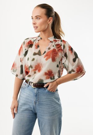 Bluse med blomster og flæsekant i gennemskinneligt stof med orange og grønne blomster på en lys baggrund, kombineret med højtaljede jeans.