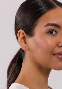 Goldene Creole mit einer Reihe kleiner klarer Steine und einer glatten Oberfläche. Getragen von einem Modell mit glattem Haar und natürlichem Makeup.