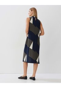 Ärmelloses Kleid in Marineblau, Khaki und weißem geometrischem Muster; hat ein Schlüsselhole-Detail am Rücken und endet knapp über dem Knöchel. Schwarz flache Schuhe getragen.