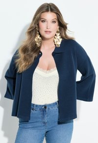 Marineblauer Strickcardigan mit dreiviertel Ärmeln, über einem weißen Oberteil mit silbernen Verzierungen getragen, kombiniert mit Jeans.
