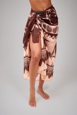 Nogi i stopy bez butów, owinięte brązowo-beżowym sarongiem w tropikalny wzór, związanym w talii, na jednolitym szarym tle.