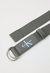 Gray fabric belt with a smooth finish, featuring a silver metal D-ring and a rectangular logo patch embossed with "Calvin Klein."