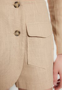 Pantaloni shorts in lino beige con due grandi tasche frontali, un design elegante e bottoni decorativi di un colore a contrasto.