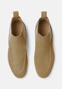 Bež suede slip-on cipele s elastičnim bočnim panelima, zaobljenim nosom i dekorativnim šavovima. Sadrži traku na peti za jednostavno obuvanje.