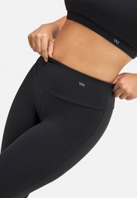 Leggings negros de cintura alta hechos de tela elástica, con una textura suave y un detalle de logo en la banda de la cintura.