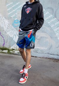 Czarna bluza z kapturem z logo koszykówki, szaro-niebieskie sportowe spodenki w wzory oraz czerwono-białe sneakersy z czarnymi sznurowadłami i białe skarpetki.