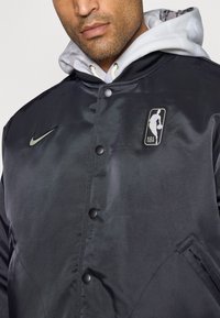 Sort satin bomberjakke med et Nike-logo og et NBA-mærke, rund sorte knapper og sidelommer. Hætteindsats er synlig.