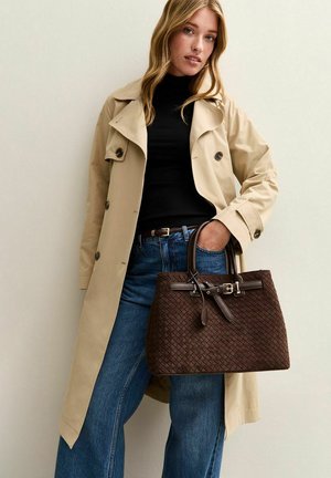 Bruine geweven handtas met een gestructureerde vorm, voorzien van een platte basis en dubbele leren handvaten, accentueert door een gesp. Draagt een beige trenchcoat.