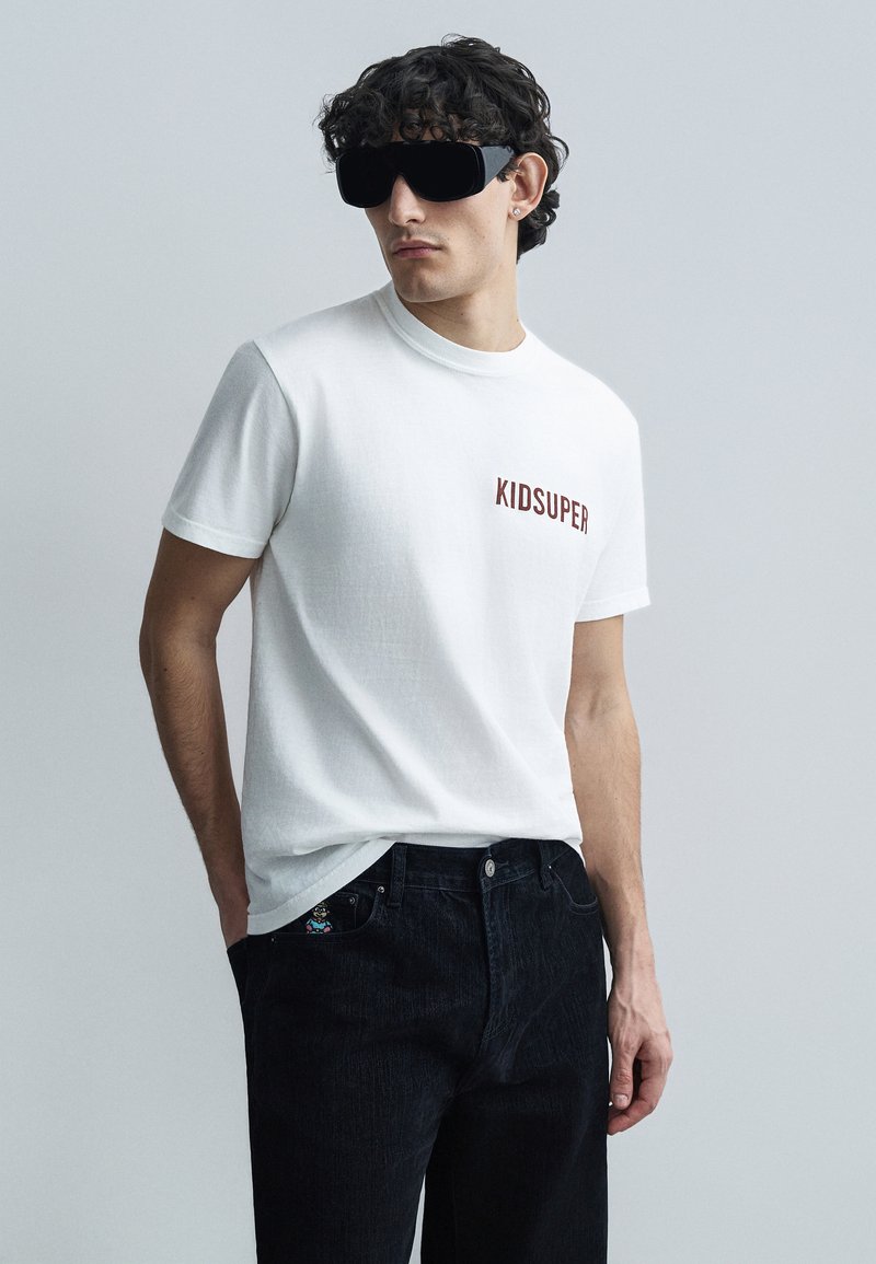 Άσπρο βαμβακερό μπλουζάκι με την επιγραφή "KIDSUPER" σε μπορντό χρώμα. Μαύρο τζιν με λεπτομέρεια τσέπης. Το μοντέλο φοράει υπερμεγέθη μαύρα γυαλιά ηλίου.