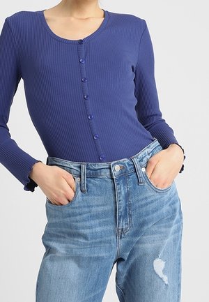 Niebieska prążkowana bluzka na długi rękaw z guzikami, zestawiona z jasnoniebieskimi, wysokim stanem, przetartymi jeansami, ręce włożone do przednich kieszeni.