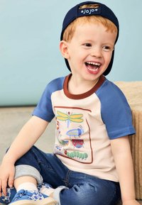 Bunt gemustertes Baumwoll-T-Shirt mit blauen Ärmeln, cremefarbenem Körper und Insektenillustrationen. Kombiniert mit blauen Jeans, weißen Socken und gemusterten Schuhen.