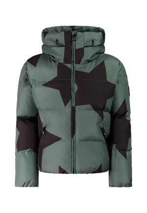 Vrouw draagt een groene donzen jas met zwarte sterren, zwarte broek, zwarte muts met ster, donkere zonnebril en zwarte schoenen, glimlachend met handen in de zakken.