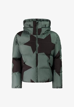 Vrouw draagt een groene donzen jas met zwarte sterren, zwarte broek, zwarte muts met ster, donkere zonnebril en zwarte schoenen, glimlachend met handen in de zakken.