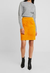 Jupe crayon en daim jaune avec une texture lisse, associée à un pull en tricot gris et des bottines noires. Présente une silhouette ajustée.