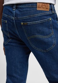 Jeans de mezclilla azul con un ajuste ceñido. Las características incluyen costuras amarillas, un parche de cuero en la parte trasera etiquetado como "Lee" y dos bolsillos traseros.