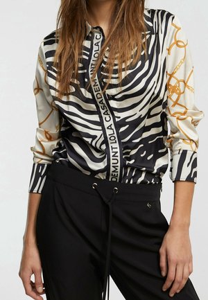 Vrouw draagt een lange mouw shirt met zebra-strepen en kettingpatroon, gecombineerd met zwarte broek met trekkoord, staand met handen ontspannen langs het lichaam.