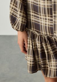 Robe à carreaux marron et beige avec taille froncée. Comprend des manches bouffantes et un ourlet à volants. Confectionnée en tissu léger.